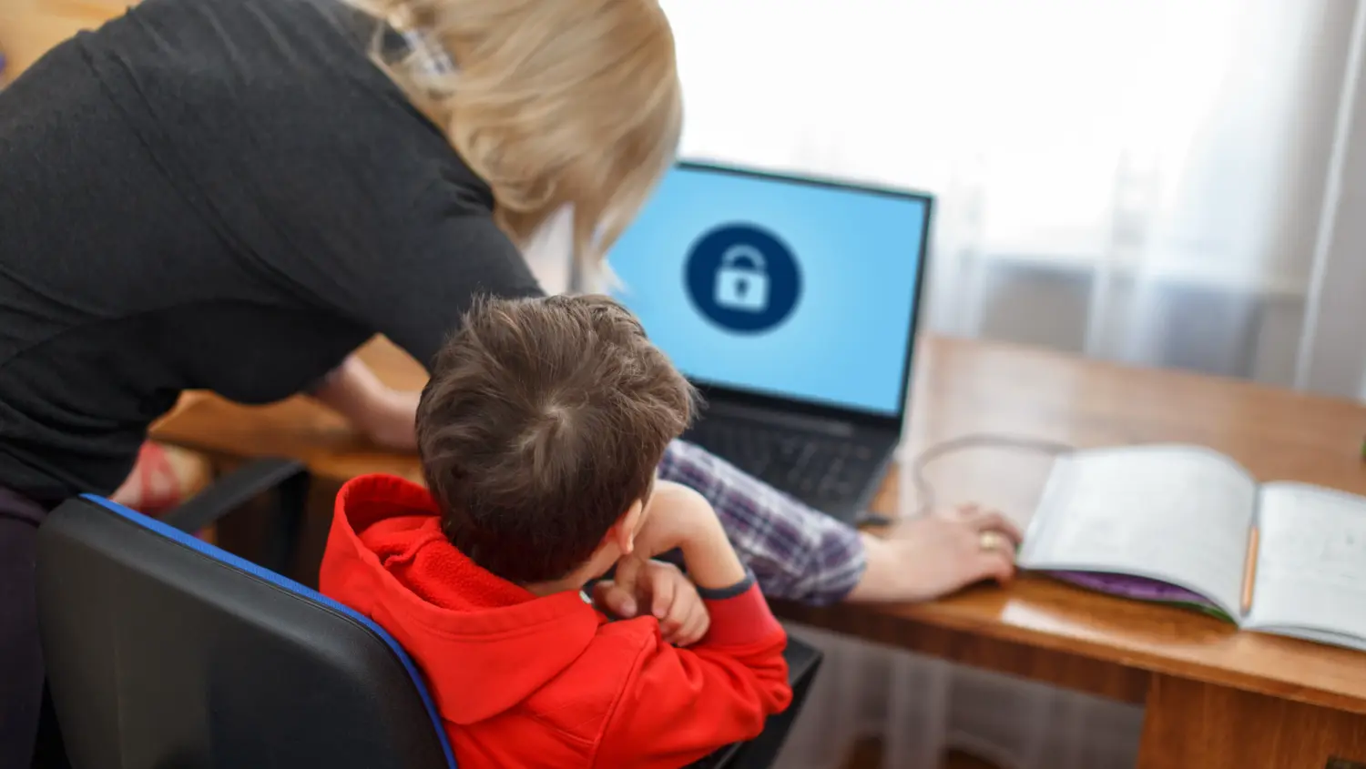 A woman helps a child using a laptop displaying a padlock icon. They are seated at a wooden desk near an open notebook, in a bright room with sheer curtains.