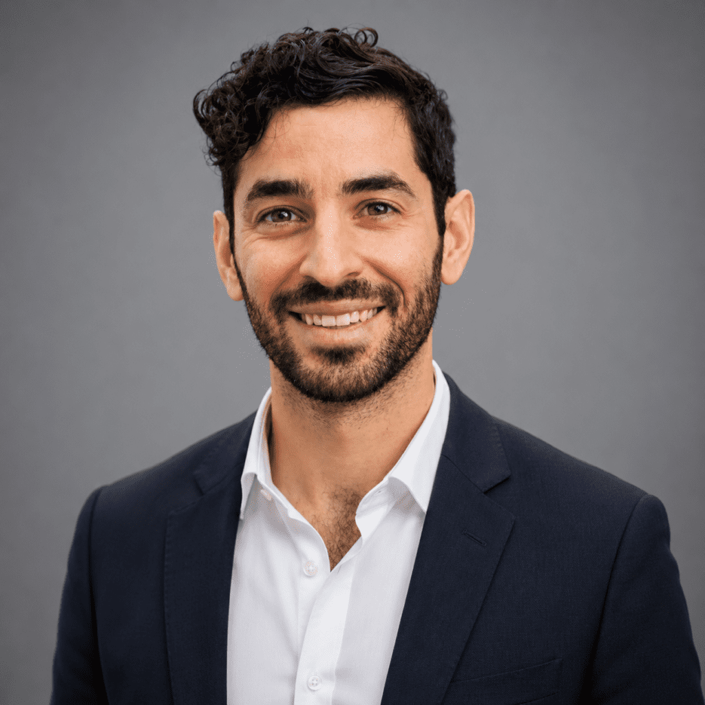 A man with dark curly hair and a beard, wearing a navy suit jacket and white shirt, smiles at the camera against a plain gray background, radiating team spirit and approachability.