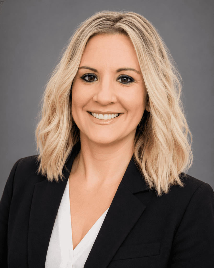 A woman with shoulder-length blonde hair wearing a black blazer and white blouse smiles at the camera against a plain gray background.