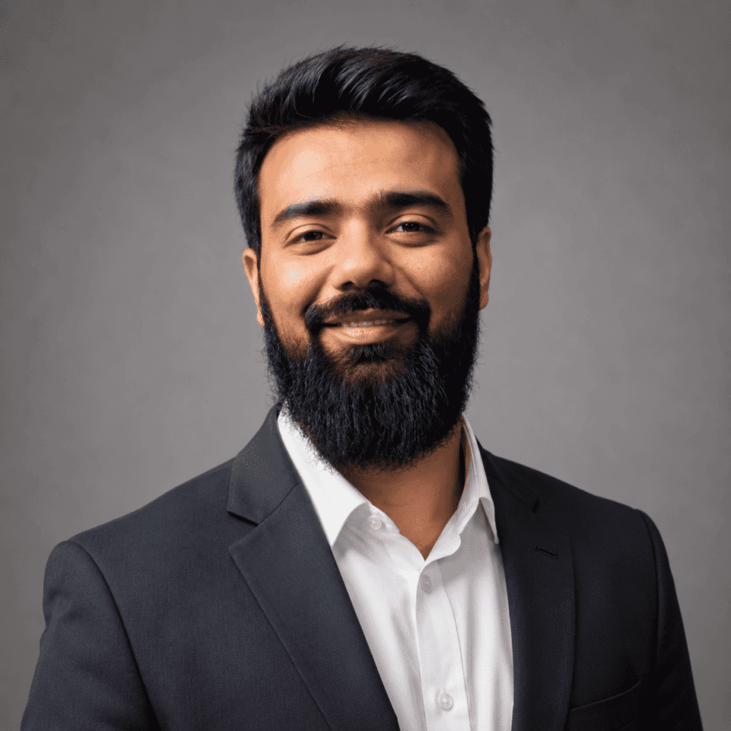 A man with a full beard and dark hair wearing a black suit jacket and white shirt smiles confidently against a plain grey background.