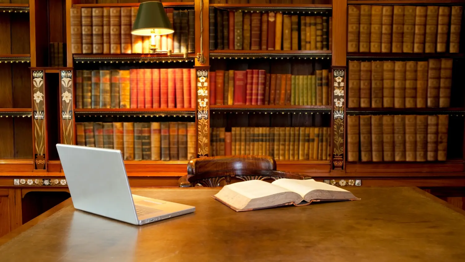 A wooden desk in a library with an open book and a laptop on it, in front of shelves filled with old, colorful books. A lit lamp is attached to the bookshelf in the background.