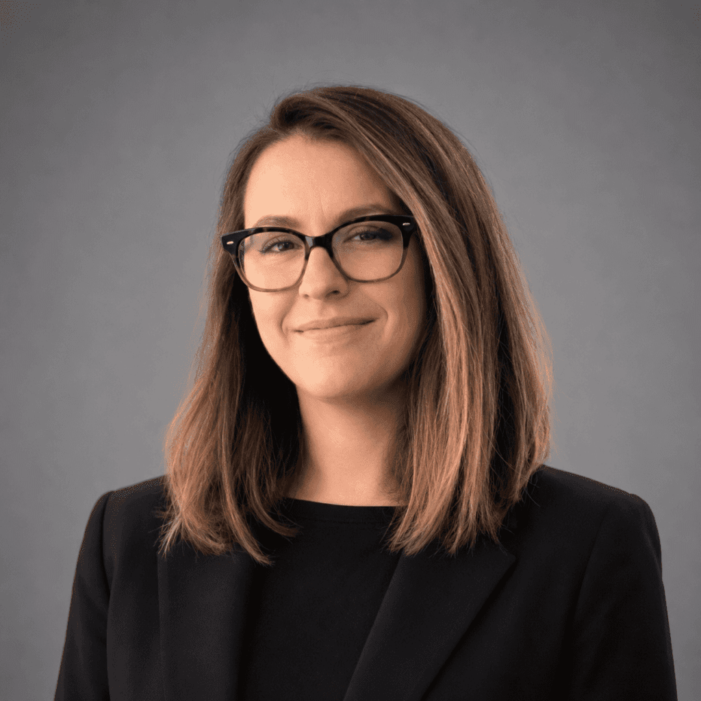 A woman with straight, shoulder-length brown hair and black glasses, wearing a black blazer and top, smiles slightly against a plain gray background.