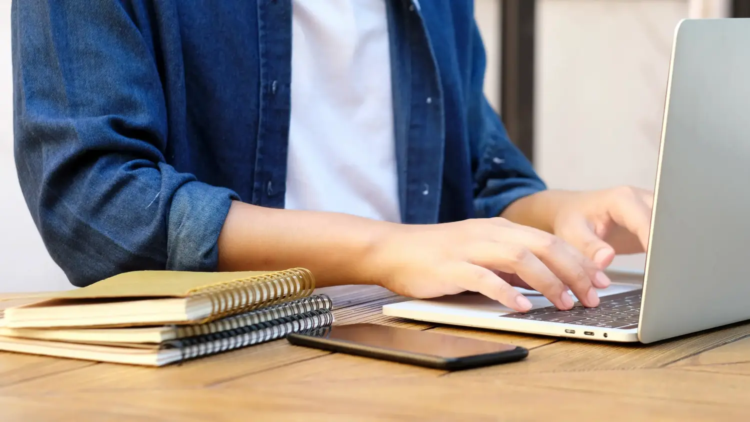 A person wearing a denim shirt is typing on a laptop at a wooden desk. Spiral notebooks and a smartphone are placed nearby on the desk. The image focuses on the person's hands and workspace.