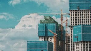 High-rise buildings under construction, covered in blue safety netting, with yellow tower cranes working around them against a bright sky with large white clouds.