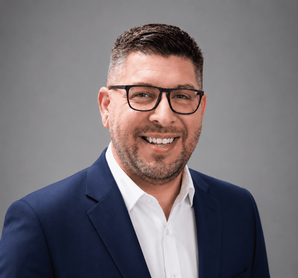 A man with short dark hair, a trimmed beard, and glasses is smiling. He is wearing a navy blue suit jacket over a white dress shirt. The background is plain and gray.