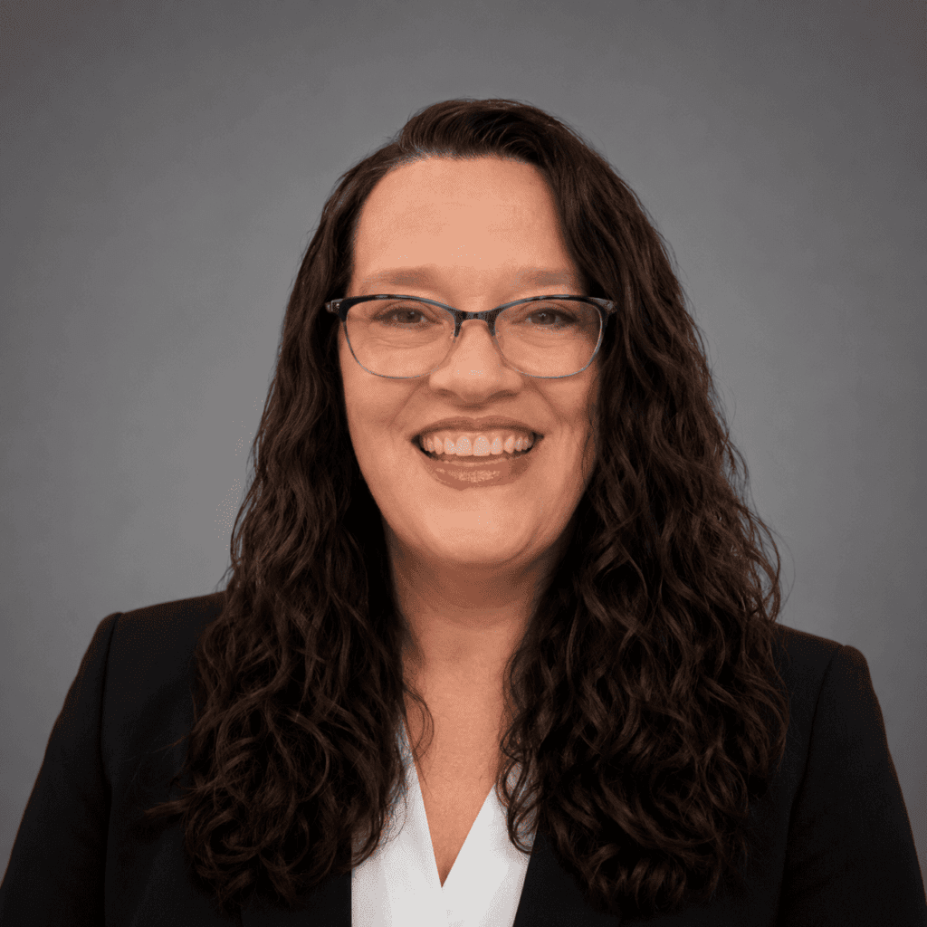 A woman with long, wavy dark hair and glasses smiles at the camera. She is wearing a dark blazer over a white blouse and stands in front of a plain gray background.