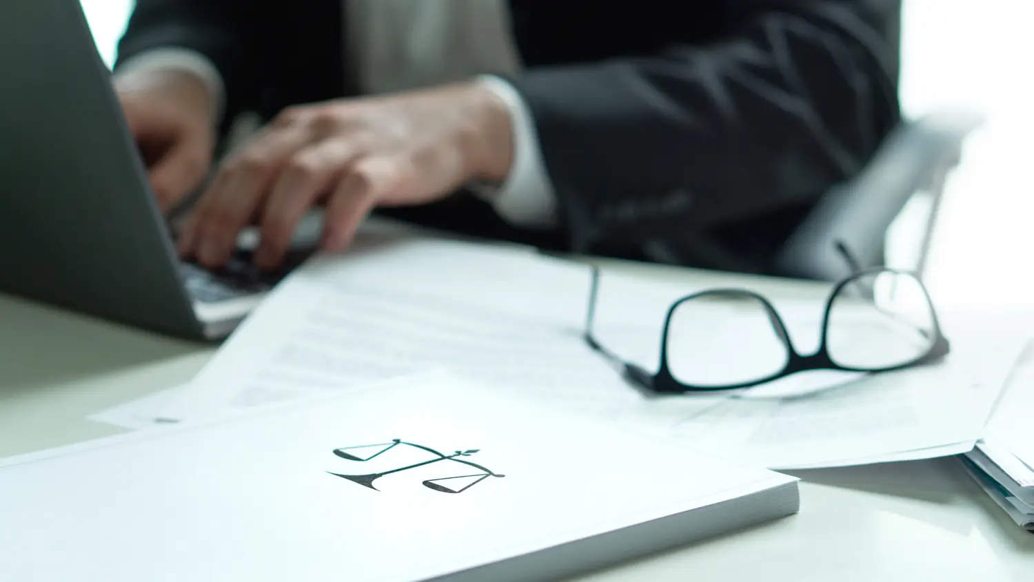 A person in a suit types on a laptop at a desk with documents, eyeglasses, and a folder featuring a scales of justice symbol, suggesting a legal or business setting.