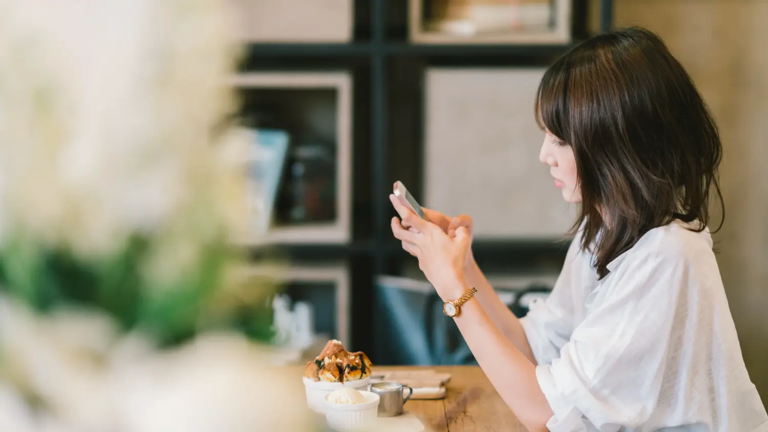 A woman with shoulder-length dark hair sits at a wooden table in a café, looking at her smartphone. There is a dessert and a small cup on the table in front of her, with shelves and blurred decor in the background.