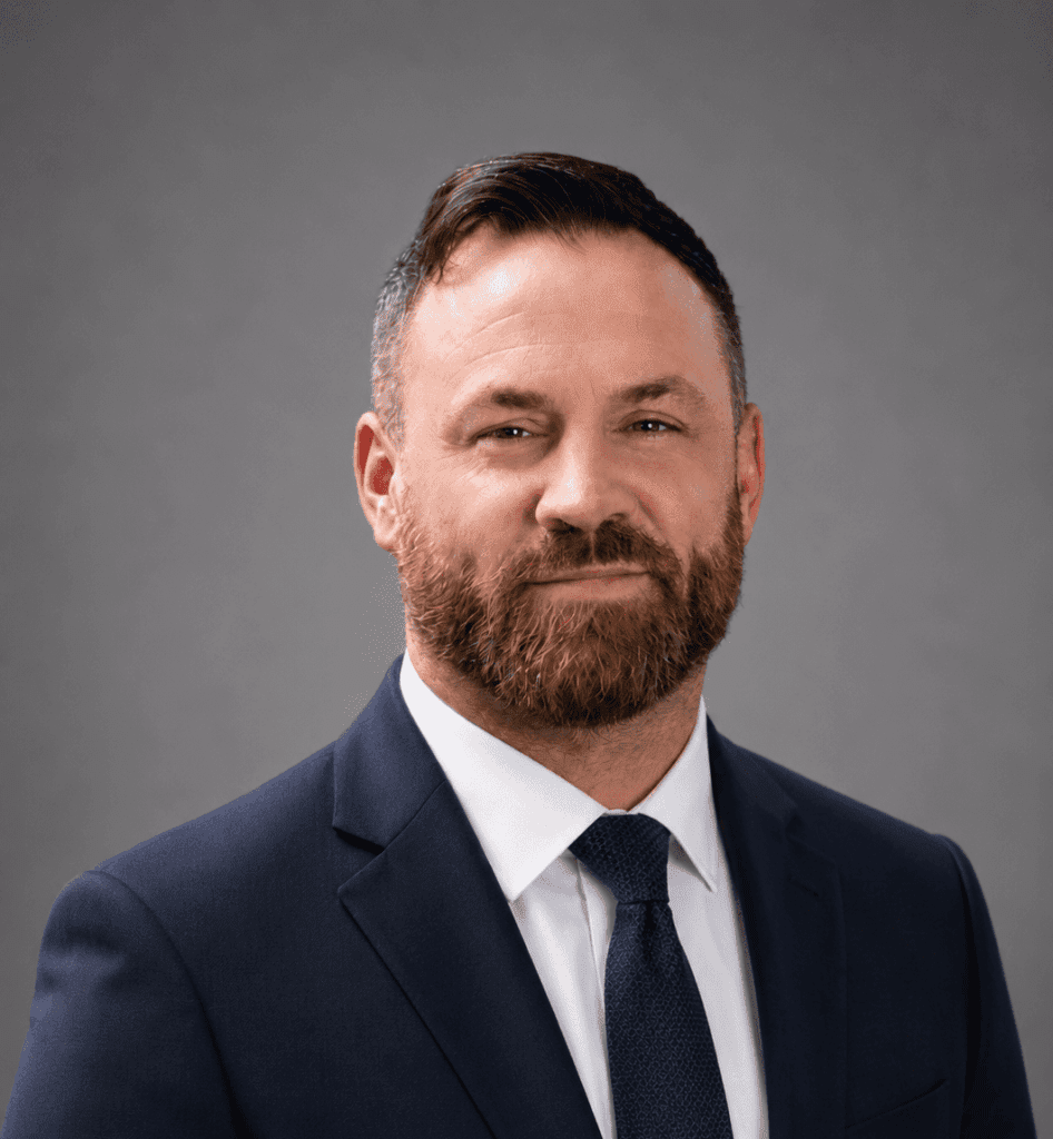 A man with short brown hair and a full beard, wearing a dark blue suit, white shirt, and dark tie, poses against a plain gray background.