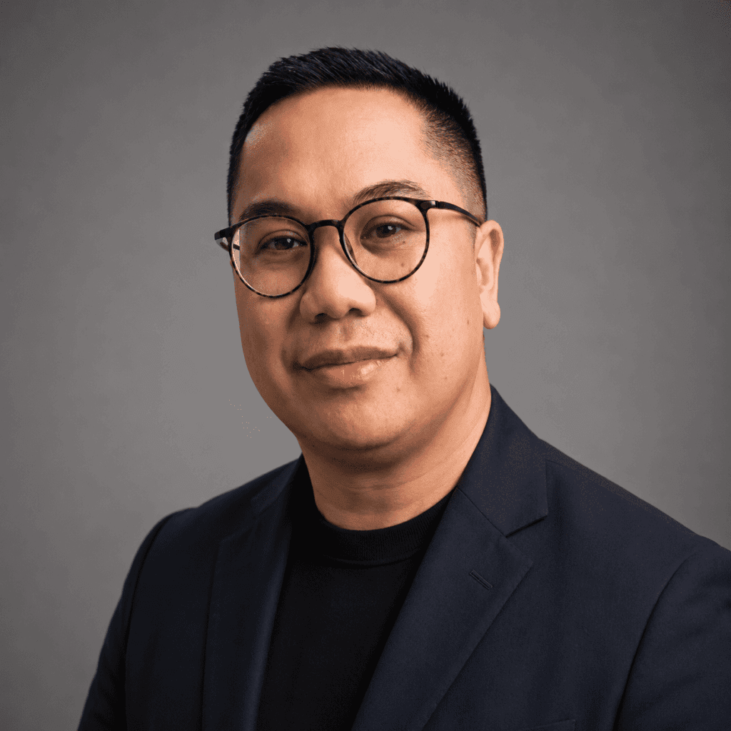 A man wearing round glasses and a dark blazer over a black shirt, posing in front of a plain, neutral gray background. He has short, neatly styled hair and a calm, confident expression.