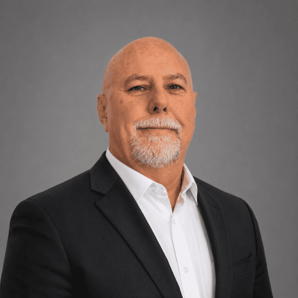 A middle-aged man with a bald head and white goatee, wearing a dark suit jacket over a white dress shirt, stands against a plain gray background, looking confidently at the camera.
