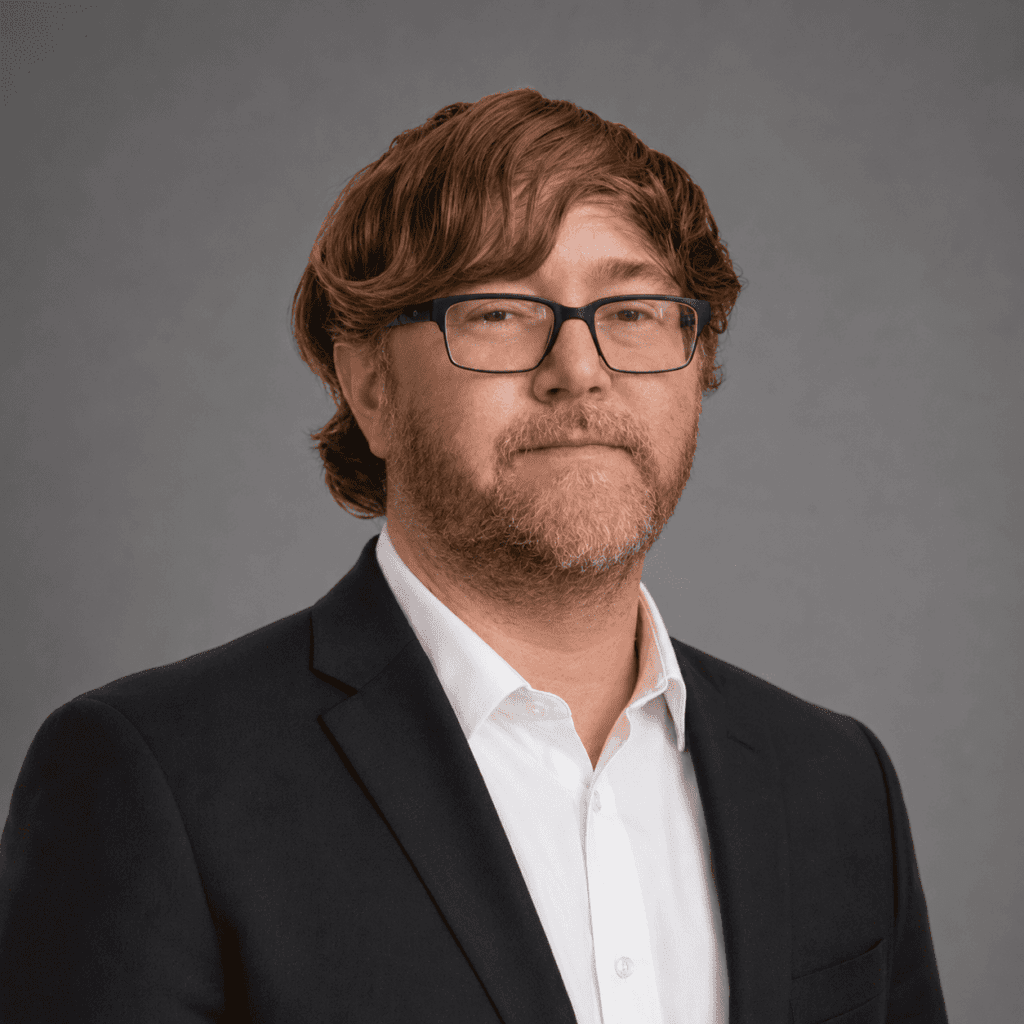 A man with light brown wavy hair, a beard, and glasses is wearing a dark suit jacket over a white dress shirt. He is posing in front of a plain gray background.