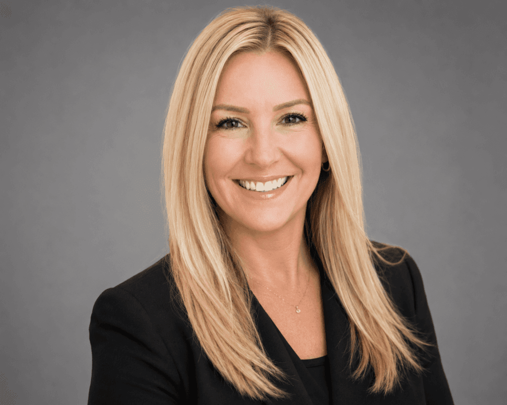A woman with long, straight blonde hair is smiling. She is wearing a black blazer over a black top and a delicate necklace. The background is plain and gray, giving the image a professional headshot appearance.