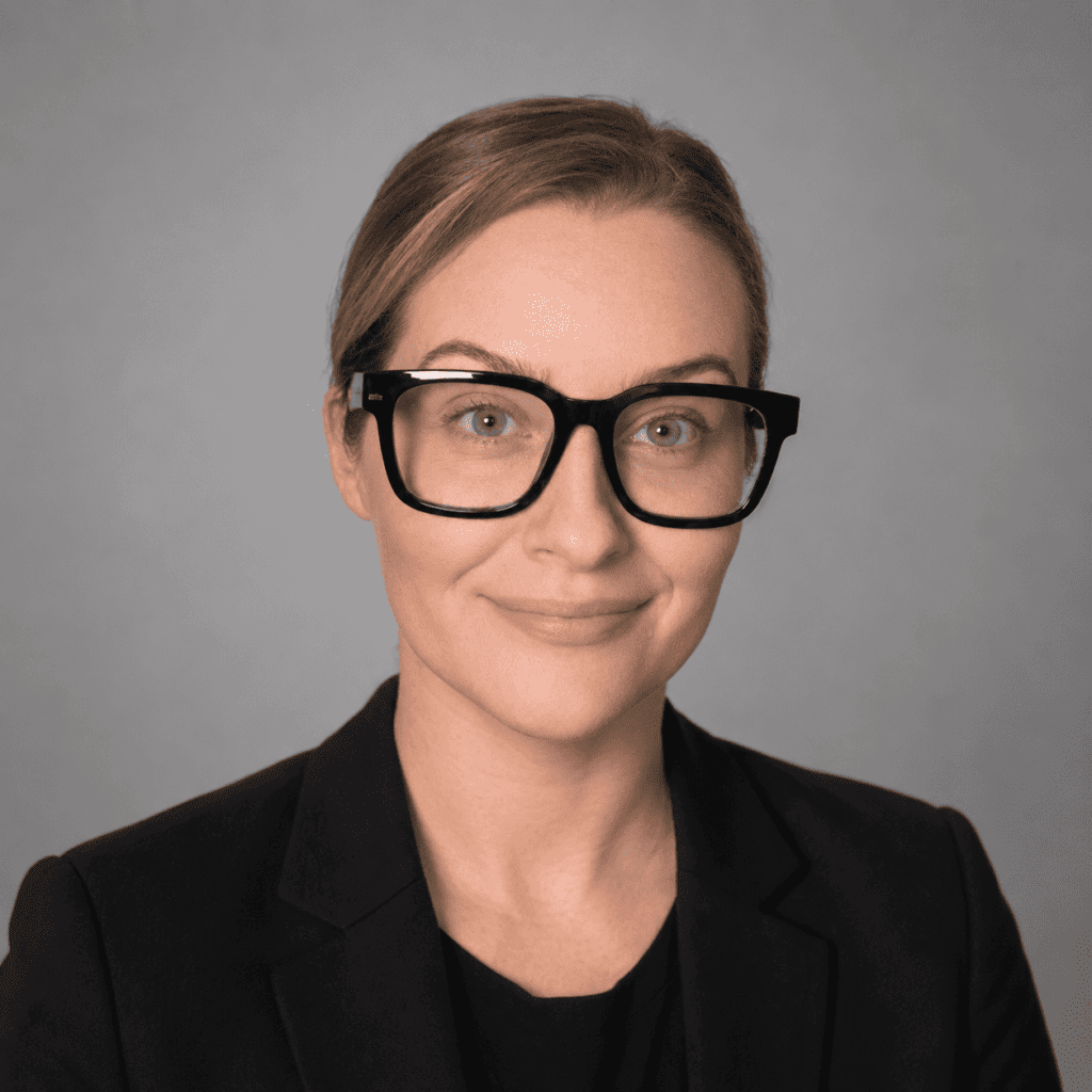 A person with light skin, wearing black glasses and a black blazer, smiles slightly in front of a plain gray background.