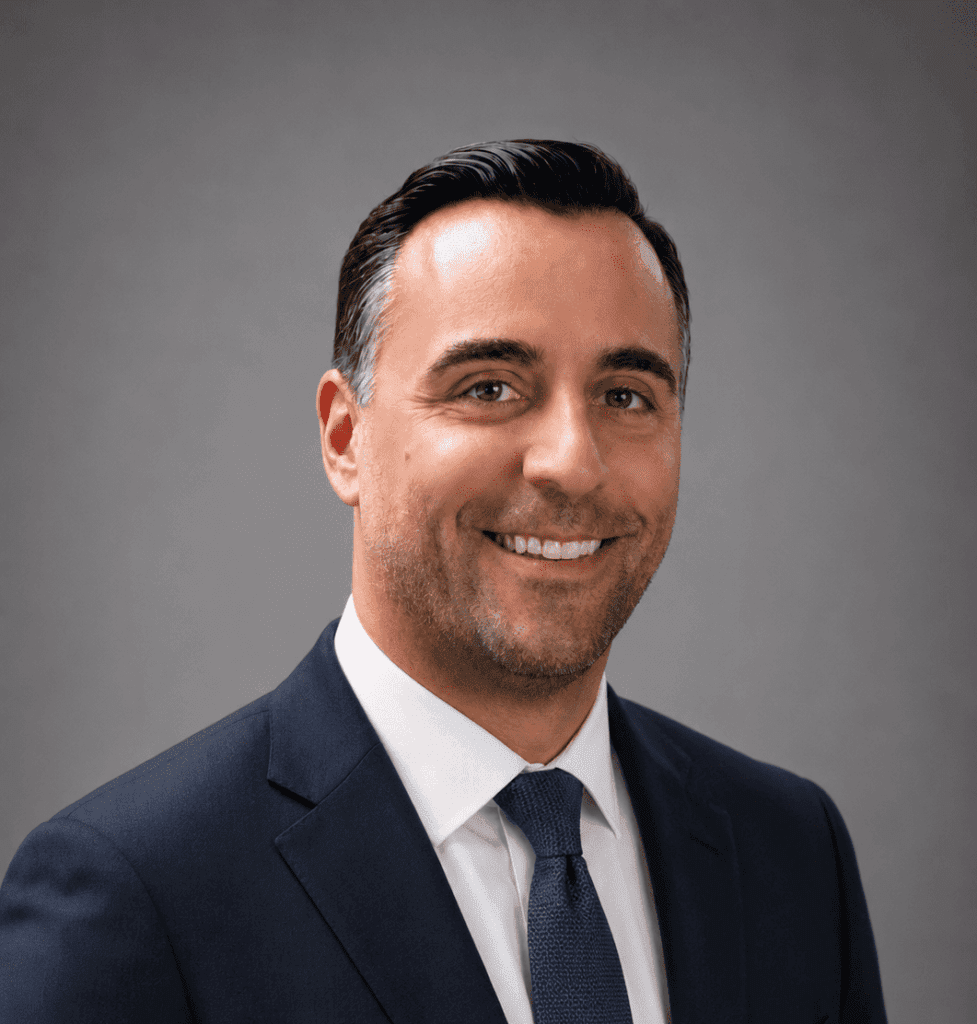 A man with short dark hair and a trimmed beard, wearing a navy suit, white shirt, and blue tie, smiles in front of a plain gray background.