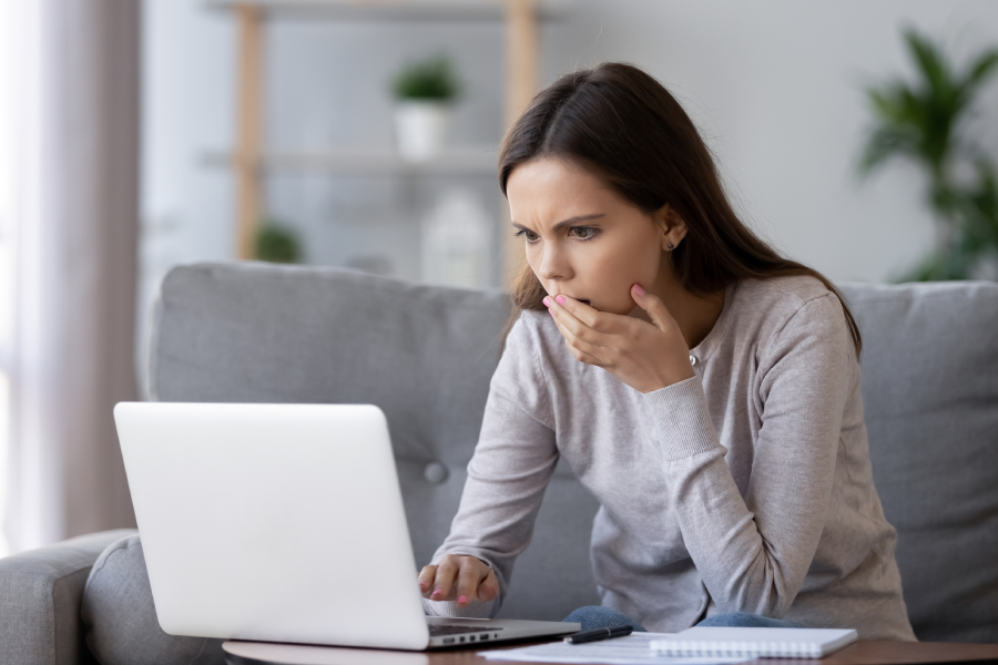 A young woman sitting on a couch looks at a laptop screen with a surprised expression, covering her mouth with one hand. A notebook and pen are on the table beside her.