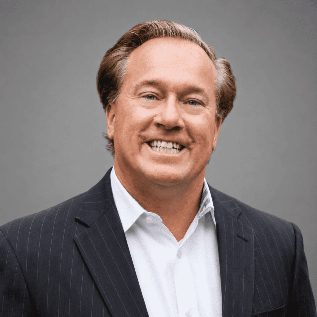 Smiling man with light brown hair in a dark pinstriped suit and white collared shirt poses in front of a plain gray background.