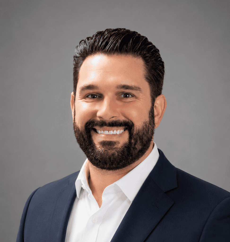 A man with dark hair and a full beard, wearing a navy suit jacket over a white shirt, smiles at the camera against a plain gray background.