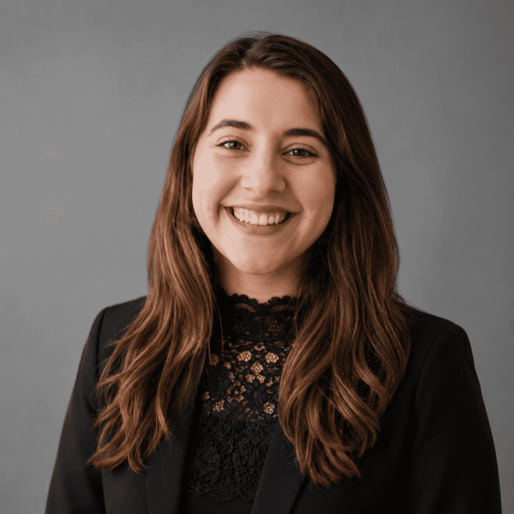 A young woman with long brown hair, wearing a black blazer and a lace top, smiles at the camera against a plain grey background.