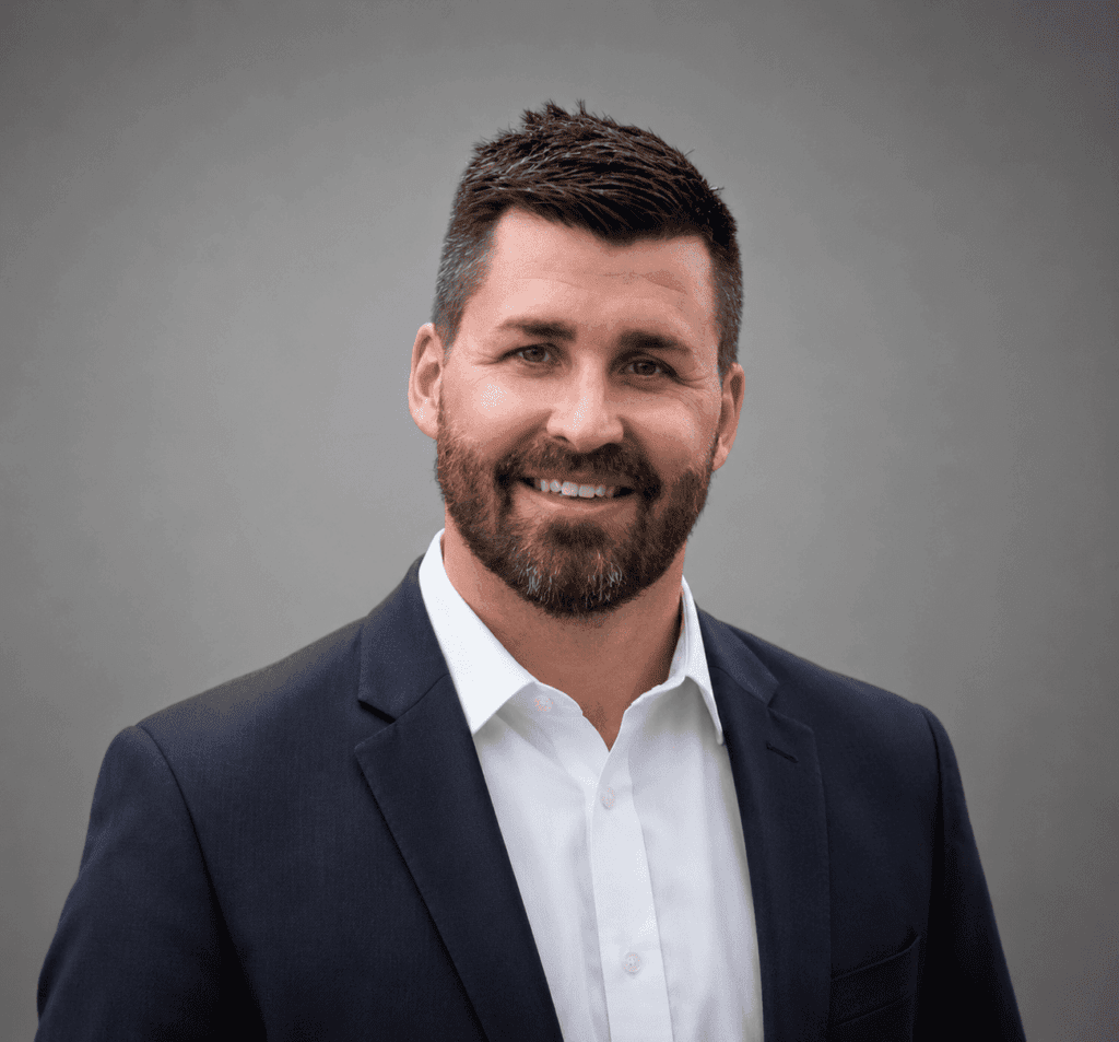 A man with short dark hair and a beard, wearing a navy suit jacket over a white shirt, smiles at the camera against a plain gray background.
