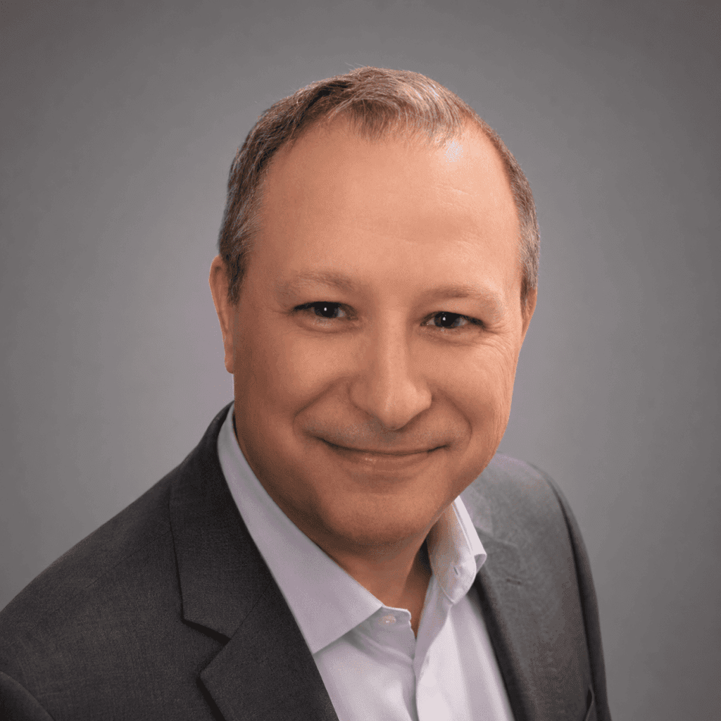 A man with short brown hair wearing a grey suit jacket and light-colored shirt smiles at the camera against a plain grey background.