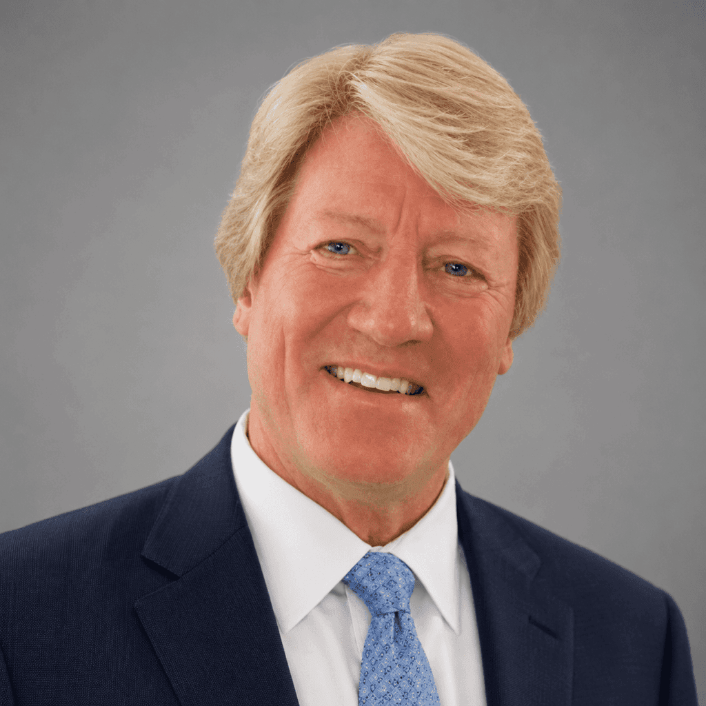 A smiling middle-aged man with blond hair wears a navy blue suit, white shirt, and light blue patterned tie, posing against a plain gray background.