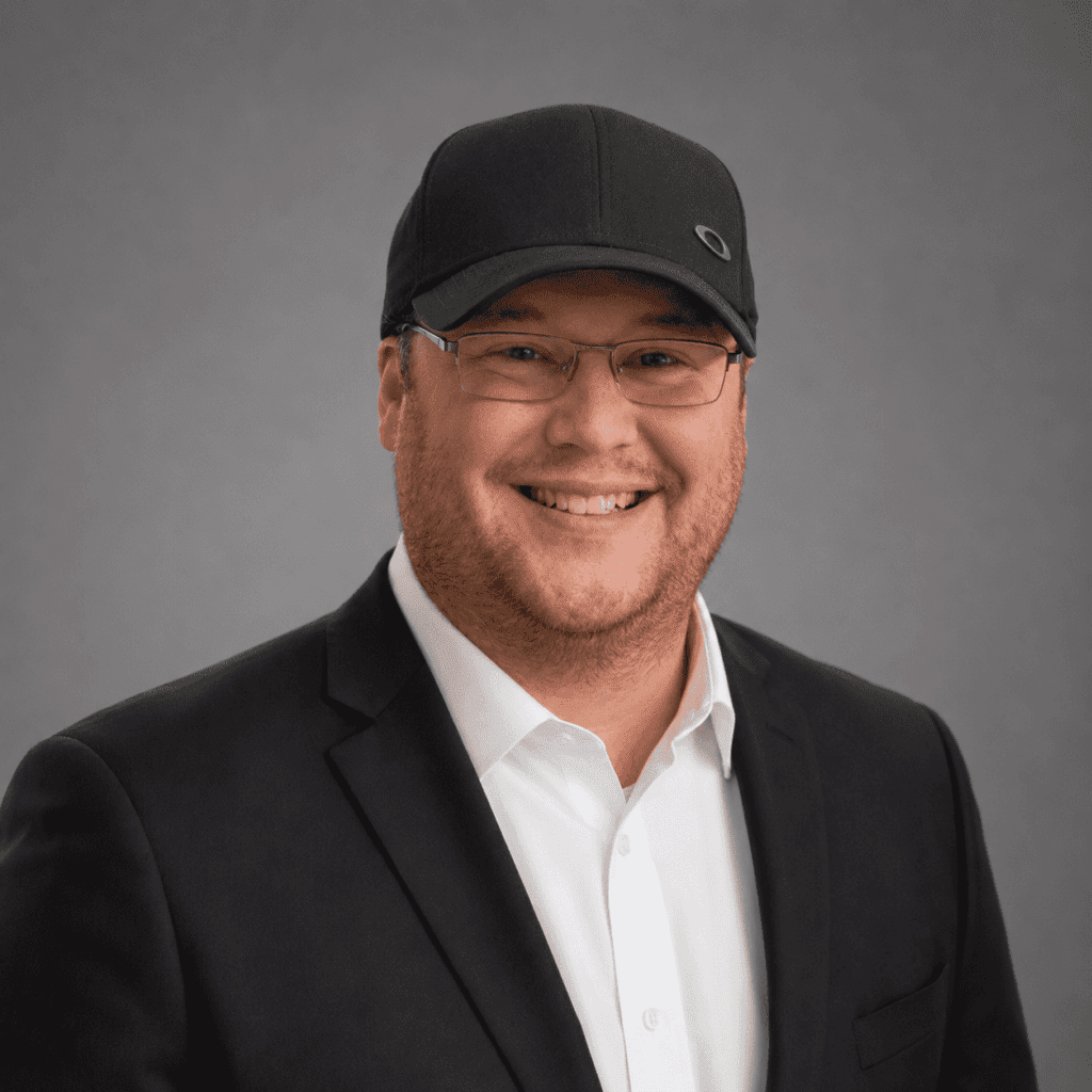 A smiling man wearing glasses, a black cap, a white shirt, and a dark suit jacket poses in front of a plain gray background.