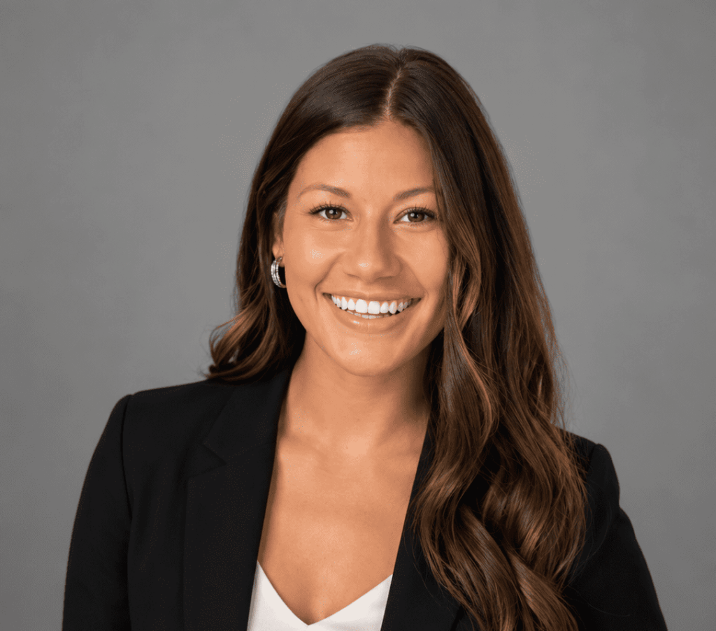 A woman with long brown hair, wearing a black blazer over a white top, smiles at the camera against a plain gray background.